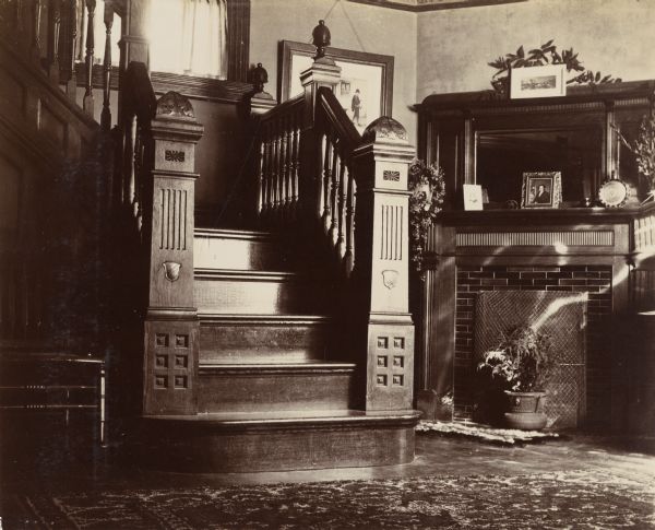 Interior view of a Victorian-era home, with a heavy, intricately carved wooden staircase. A brick fireplace with a mirrored mantel adorns the adjoining wall on the right.