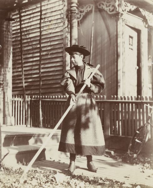 A young woman wearing a dress and a hat rakes leaves in a yard. In the background, a neighboring house is adorned with detailed fretwork brackets on an outdoor porch.