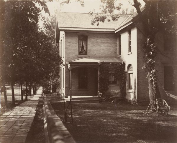 An exterior view of a two-story brick house and its surrounding yard. The photograph is taken looking down the street and sidewalk. The house boasts a small, outdoor porch with modest spandrels and dental work. A hammock is visible in the yard, along with a wooden chair and croquet mallets.