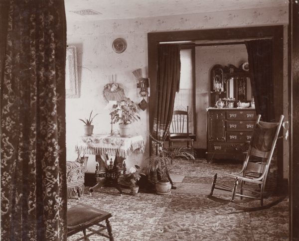 Interior view through curtained doorway of the Woodard's Victorian-era home. Elaborate rugs and wallpaper adorn the house, in addition to plants and wooden furniture.
