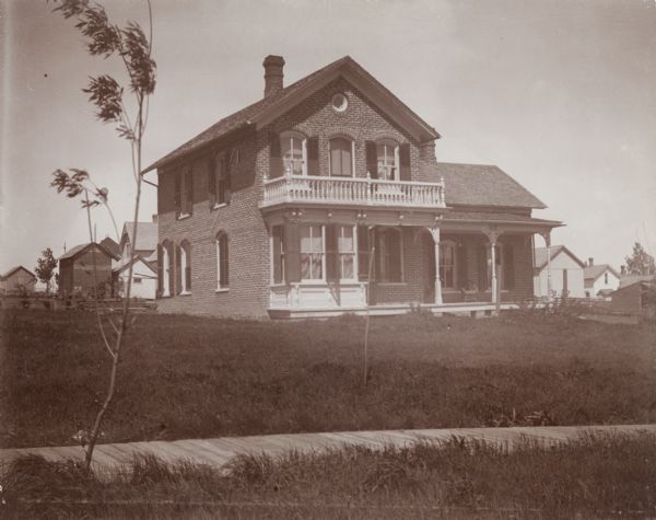 Exterior view of the Woodard home. The two-story house is made of brick and has black shutters framing the windows. There is a first and second story outdoor porch with white balusters, decorative brackets and corbels.
