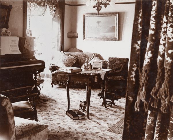 Interior view of the Woodard home. Looking through curtained doorway into the parlor, is a piano, small sofa, conversation table and several chairs. Various decorative details adorn the room such as flowered curtains, paintings, and a ceramic dog.