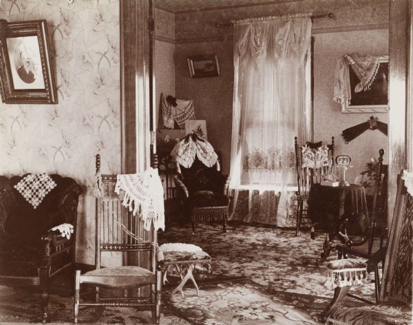 Interior view of home of Chas Cornelius, the Register of Deeds. The house is adorned with elaborate carpeting and decorative wallpaper. Additionally, various tapestries, paintings, pictures and wooden furniture fill the room.