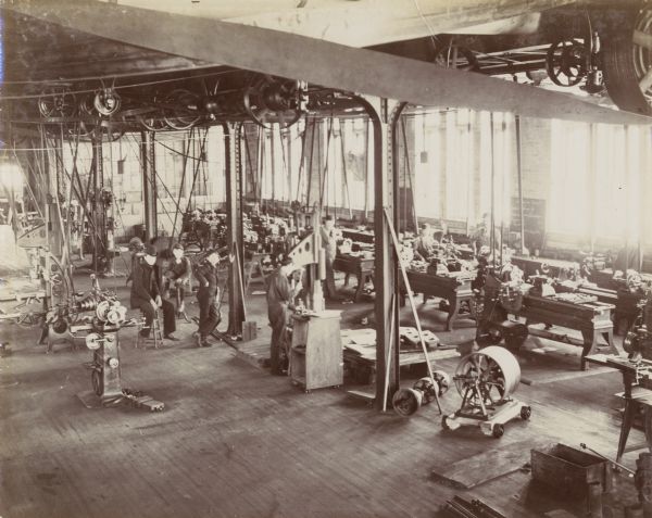 Elevated view of interior of the University of Wisconsin-Madison's Machine Hall. Several men work in the large, warehouse-like work space amidst numerous machines. The work space is brightly illuminated by the sun shining through a wall of windows.