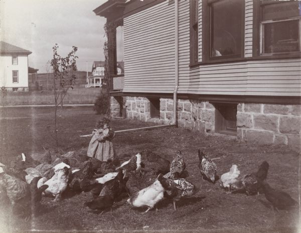 A young girl feeds a flock of chickens outside the Leslie Bird house. Several other residences are visible in the background.