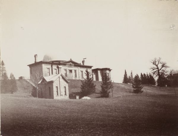 Exterior view of the University of Wisconsin-Madison observatory. The building stands on top of a hill and is surrounded by several trees. In the foreground is a smaller building at the foot of a flight of steps on the hill.