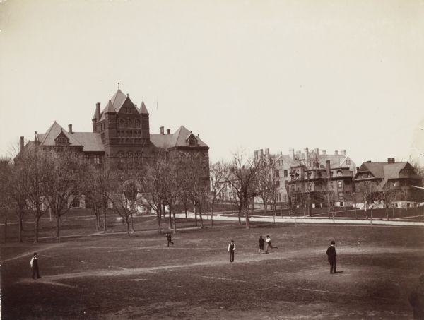 Exterior view across field of the University of Wisconsin-Madison Science Hall. A group of men appear to be playing baseball on the open lawn along Langdon Street. In the background are several homes and university buildings.