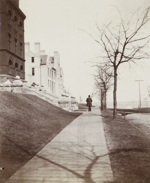 A man walks toward Lake Mendota on a wooden sidewalk in front of Science Hall on the University of Wisconsin-Madison campus. The Chemical Laboratory building is behind Science Hall on the left.