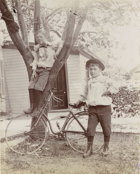 Two boys, Verne and Fay Canon, pose outdoors. Verne sits in a small tree and rests his feet on the front tire of a bicycle, while Fay stands and rests his hand on the bicycle seat. A stack of lumber and an outbuilding are visible in the background.
