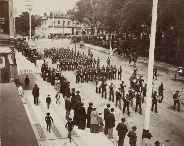 An elevated view of a Decoration Day parade, near what appears to be Capitol Park. A small crowd gathers at the edge of the street to view a military band and soldiers marching.