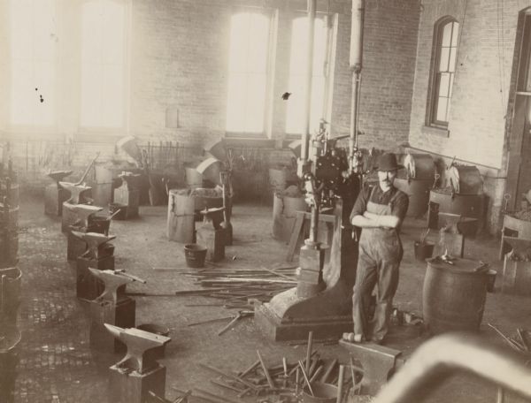 The interior of the University of Wisconsin-Madison blacksmith shop. A man stands with his hands crossed in the shop, surrounded by various blacksmith implements, including anvils.