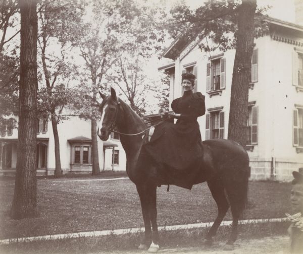 Mrs. Sterling, an instructor in German at the University of Wisconsin-Madison, rides horseback in a neighborhood. In the background are several residences, and in the right foreground is a child wearing a hat. The house in the background to the right is at 811 State Street.