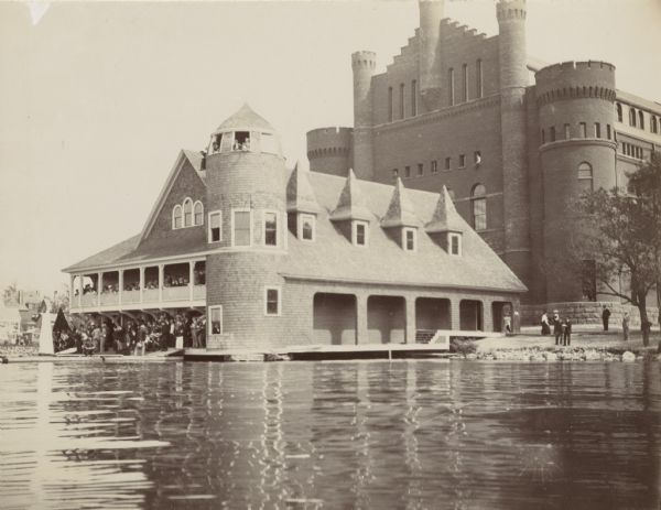 View from Lake Mendota of the University of Wisconsin-Madison boathouse. A large crowd gathers on the porch and landing of the boathouse, perhaps to watch a rowing regatta. The University of Wisconsin-Madison Gymnasium (Old Red or Red Gym) is behind the boathouse.