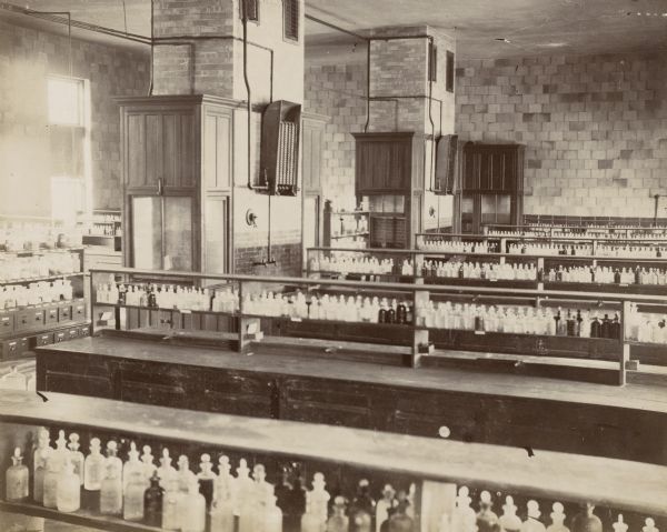 The interior of the University of Wisconsin-Madison Chemical Laboratory. Rows of glass bottles line the tables and two fume hoods are installed on the brick pillars.