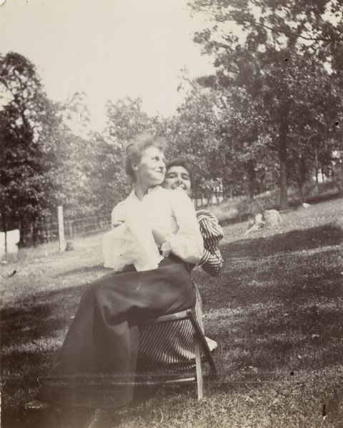 Two women pose for an outdoor photograph at the Jonas' campsite at Merrill Springs. One woman sits on a stool with her needlework while another kneels behind her and peers over her shoulder.