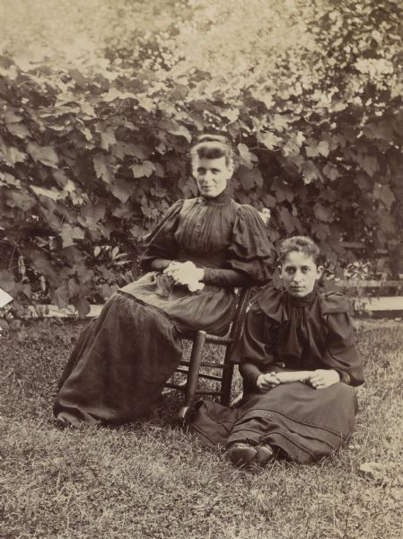 Two women sitting in yard. One woman sits in a rocking chair, the other woman sits on the ground, holding an ear of corn in her lap. In the background a vine grows along a fence.