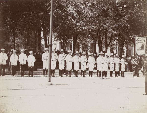A group of "Modern Woodmen of America" stand at attention in a parade. The men are wearing uniforms that say, "MWA 51," and holding axes in their hands. A man on the right holds a banner.
