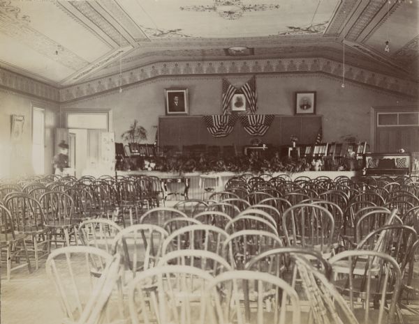 Interior view of the chapel at the Wisconsin School for the Deaf. Rows of empty, bow back chairs face a stage, where more chairs are set up under bunting and flags. A woman is near the doorway on the left.