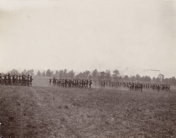 Formations of soldiers at Camp Douglas march across a large parade field. A line of trees are in the distance, and a building with a chimney is visible above the trees on the right.