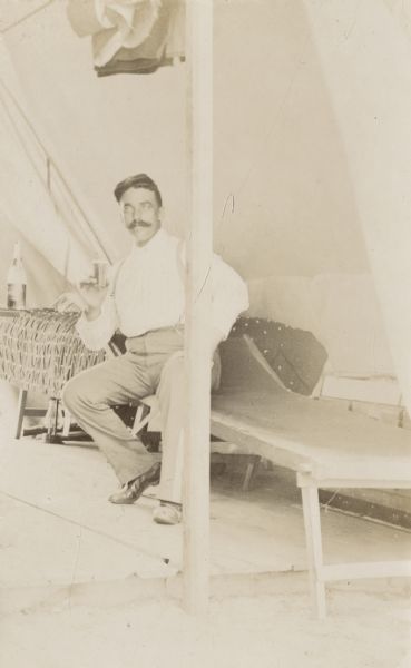 Major Joachim sits on a cot in his tent holding a drinking glass.