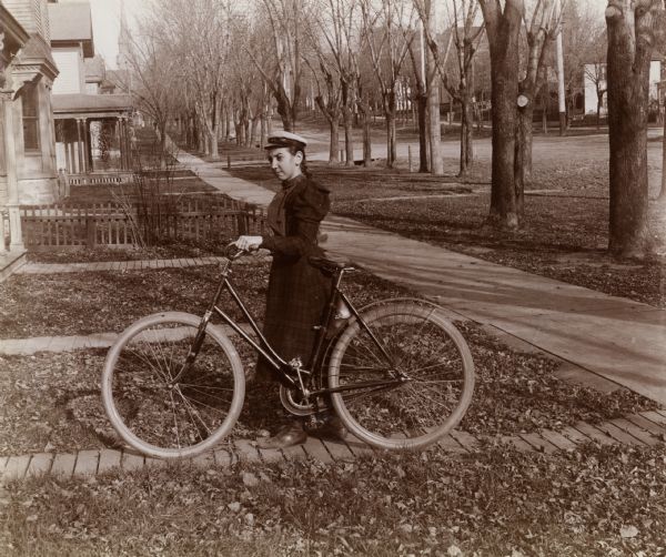 Mrs. Annie Coon stands in a front yard with a bicycle, probably on the 400 block of West Washington Avenue. A long line of trees grow along the street of the residential neighborhood. The First Congregational Church is visible in the background at 222 West Washington Avenue.