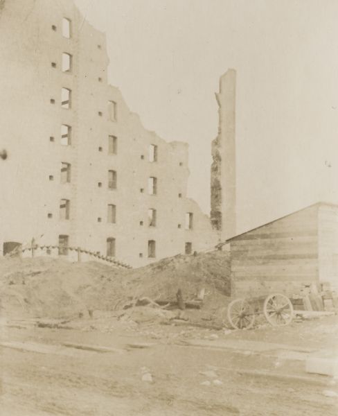 View of the ruins from a fire in the 3rd Ward. A section of a tall building, perhaps a wall, still stands. In the foreground on the right is a cart near a wood shed.