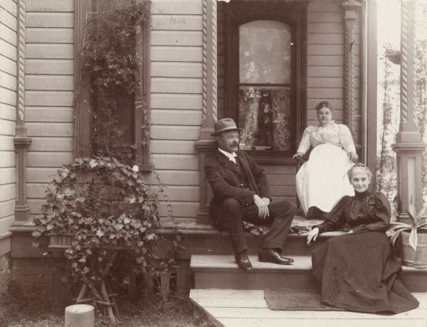 Mr. and Mrs. Chas Joss and Mrs. Van Dyke sit on a residential porch. A planter of ivy is in front of a narrow window on the left, and the porch has decorative, turned posts.