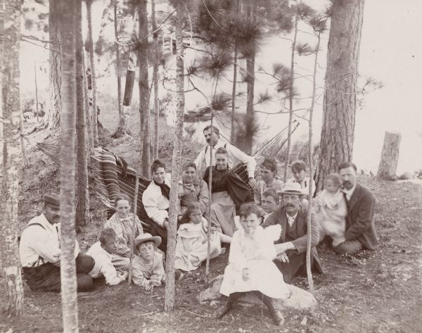 A group of men, women, and children pose for a photograph as they relax in the woods around a large hammock. In the background is a shoreline.