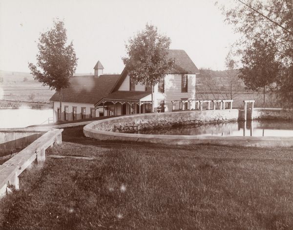 Exterior view of the Wisconsin State Fish Hatchery (aka Nevin Fish Hatchery, 3911 Fish Hatchery Rd). A salt-box house with an additional wing stands next to several reservoirs. The house has a small, decorative porch with small brackets and simple posts.