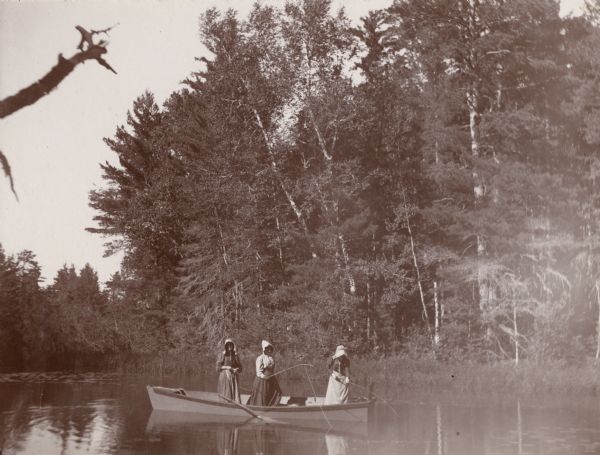 Three women, wearing dresses and bonnets, standing up in a small boat fishing. A tree-lined shore is visible in the background.