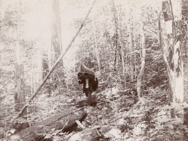 A man carries a canoe on his shoulders while portaging through a thickly-wooded area.