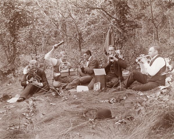 Five men are sitting on the ground in the grass playing various horned musical instruments. The corresponding caption reads: "Picnic at Winnequah." They are reading from music set up on music stands.