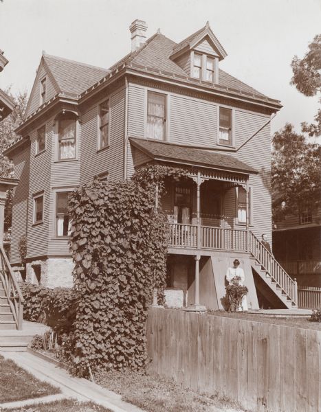 Exterior view of Judge A.S. Zimmerman's home on West Main Street. Mrs. Zimmerman stands outside the house in the yard and tends to a plant. Mr. Zimmerman stands on the stairs to the porch.
