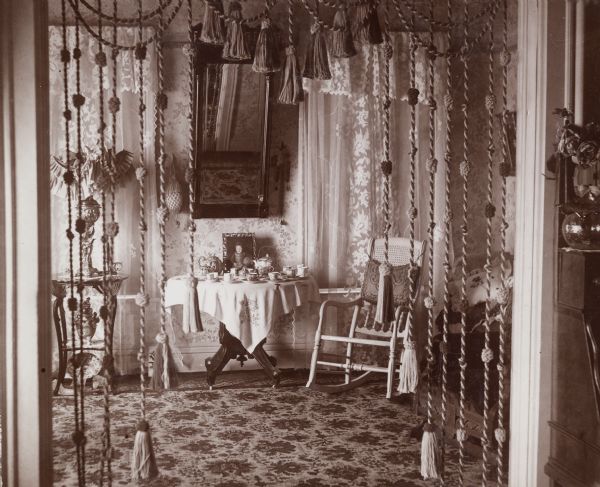 Interior view through tasseled doorway of a room in the James Nevin home. Nevin was the supervisor of the State Fish Hatchery. The room is decorated with mirrors, tables and chairs. Two teapots, with cups and saucers, are set up on the table.