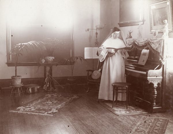 A nun stands near a piano and plays a lute-like instrument in a house on West Washington Avenue.