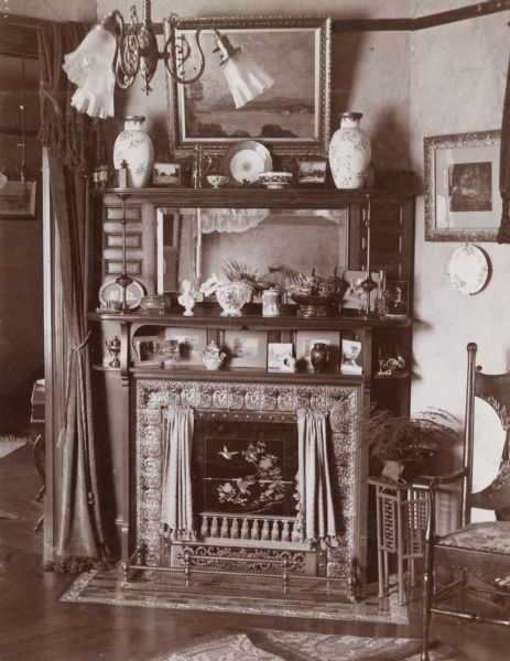 An interior view of the Eugene Eigheny home at 241 Langdon Street. Decorative glass with painted birds is visible in the fireplace opening. There is a large mantel with beveled glass. Various porcelain vases, miniature busts and tableware pieces are displayed on and around the mantel.