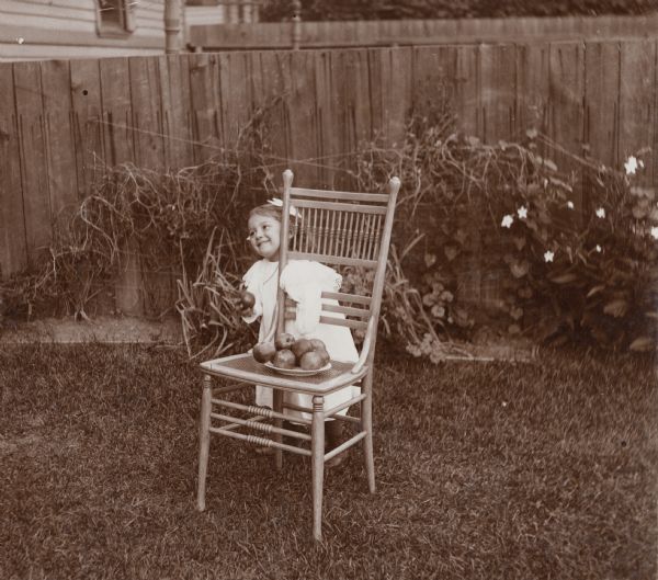 A young girl, Helen Phillips, stands in a yard near a fence behind a wooden chair. She reaches though the decorative panels on the back of the chair and grabs an apple from a plate that rests on the caned-seat.