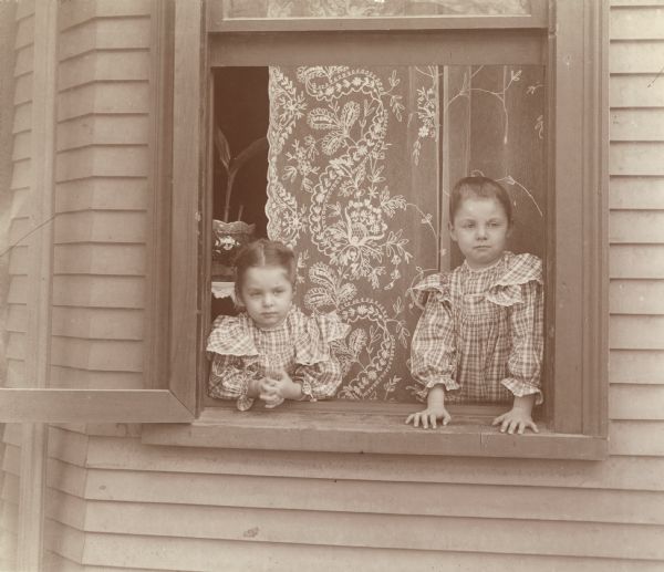Two young children look out a window at the F.D. Eyerly residence. The children are dressed in matching, plaid-patterned dresses and stand in front of a lace curtain.