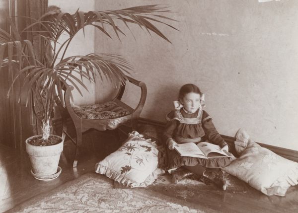 A young girl, Dorothy Hubbard, sits on a hardwood floor surrounded by pillows and reads a book. Next to the girl a large house plant grows in a pot, and a curved bench rests in the corner.