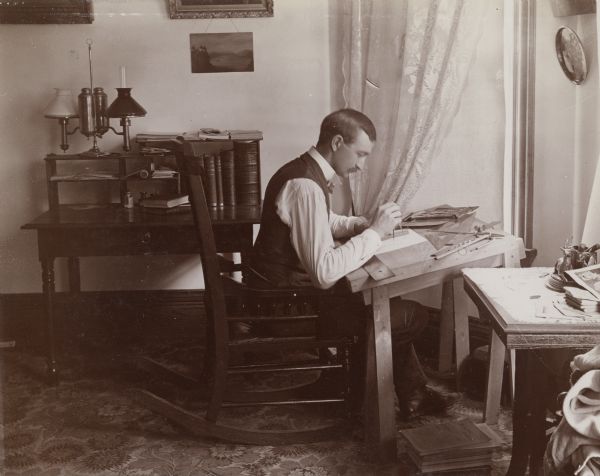 Mr. Edward Schildhauer sits in a rocking chair at an angled architect desk. The desk is covered with papers and various drafting tools such as a protractor, compass and ruler.