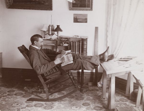 Mr. Edward Schildhauer relaxes in a rocking chair with his feet on a desk. He is reading a newspaper.
