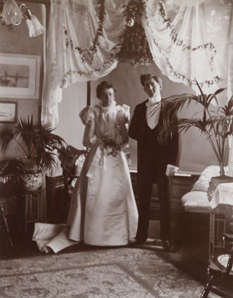 Mr. and Mrs. D.W. Osborn stand in a Victorian-era home for their wedding portrait. Shades are pulled over a bay window in the background and white fabric decorated with plants is draped from the ceiling archway to create an aesthetically-pleasing backdrop.