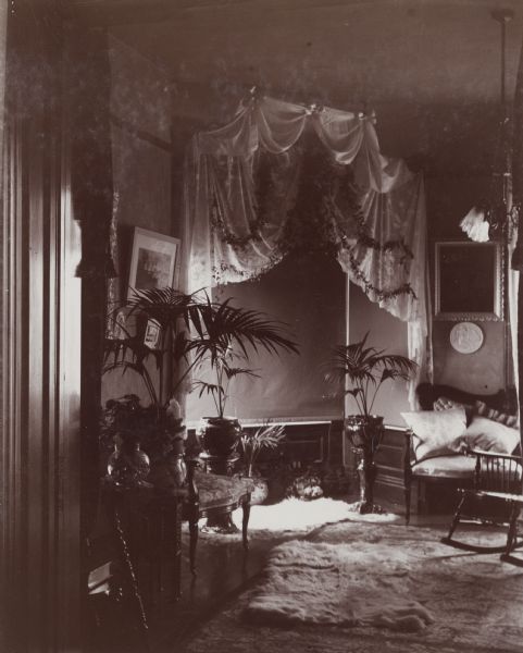 Interior view of a decorated bay window in a Victorian-era home. The window area was used as a photography back-drop so the shades are drawn and fabric is draped over the opening to the bay window area. There are decorative potted plants and rugs throughout the room.