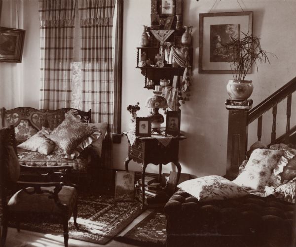 An interior view of a sitting room in the W.B. Hathaway house on West Johnson Street. The Victorian-era room consists of two plush pieces of furniture: a loveseat and a daybed. Additionally, a wooden chair, end-table and decorative shelving unit are visible. Amidst the furniture are various framed pictures, a plant, a porcelain lamp and decorative area rugs. A staircase is on the right behind the daybed.