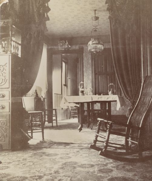 Interior view of the H. Schildhauer home at 13 North Webster Street. The photograph was taken looking from the parlor into the dining area. The Victorian-style home has patterned wallpaper and rugs, heavy draperies, elaborate glass light fixtures and various pieces of wooden furniture.