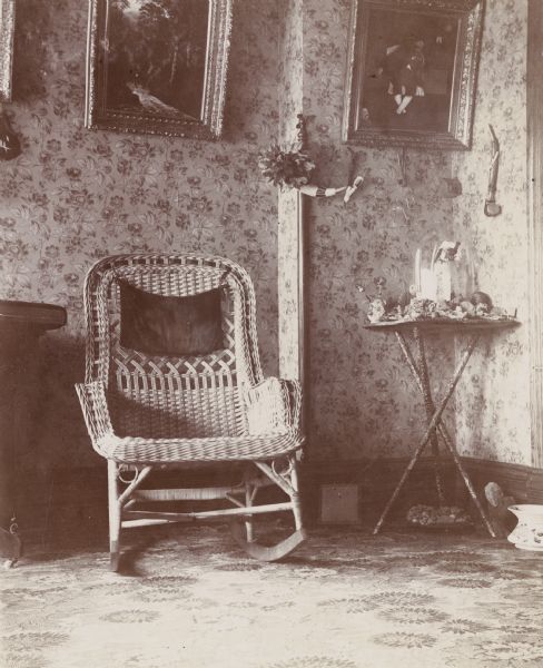 An interior view of a sitting room in the H. Schildhauer residence at 13 North Webster Street. Several paintings hang on the wall behind a wicker rocking chair, and a small tripod table resides in the corner of the room, on which a glass dome is surrounded by shells and pine cones.
