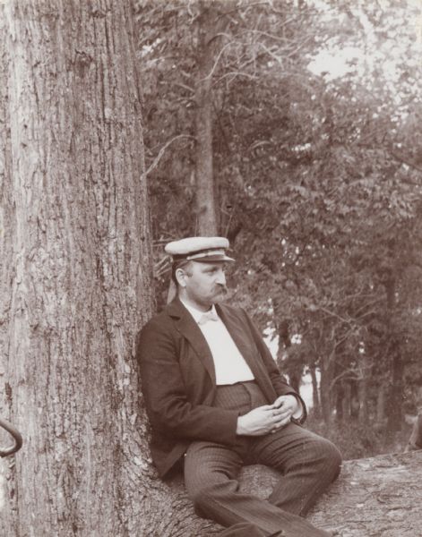 Mr. H. Schildhauer sits at the base of a large tree. He wears pinstripe pants with a jacket, a modest bow-tie and a cap.