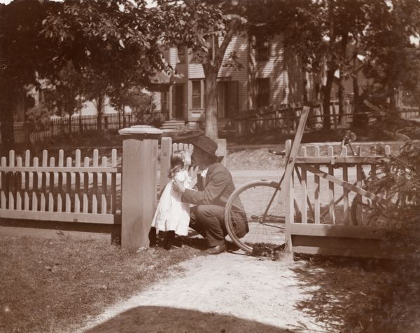 F.D. Eyerley crouches down to give his daughter a kiss. A bicycle is propped against a wooden fence which runs the perimeter of the front yard of the West Johnson Street home.