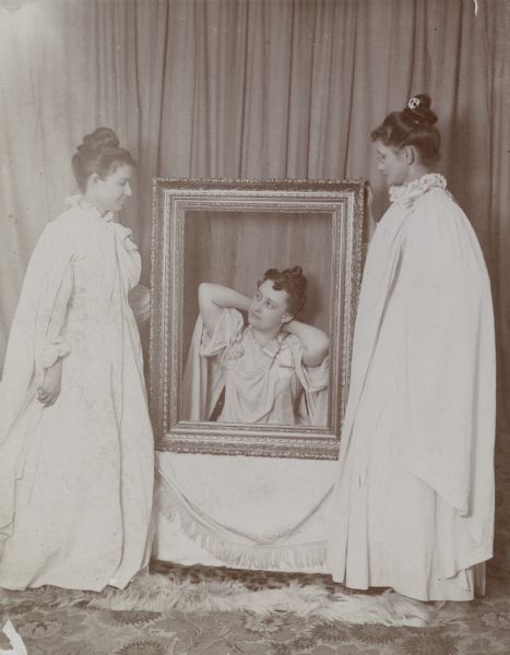 Three women, from left to right: Mrs. Vandercook, Mrs. Osborn and Mrs. Schildhauer, pose in white Grecian-inspired costumes. Mrs. Osborn sits behind a large, empty picture frame while the other two women stand.