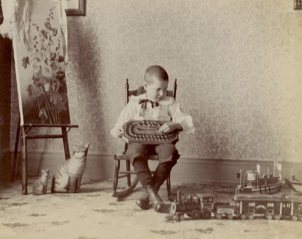 Young boy, Philip Mills, sitting in chair holding a board game. A toy train and toy boat are on the floor. On the left a painting is on an easel, and two fabric cats are positioned underneath.